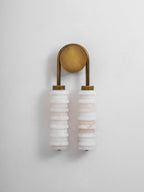 Small unlit Clérachic sconce in antique brass on a pale wall, two white alabaster columns hanging beneath the round plate.