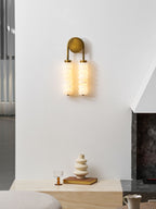 Clérachic antique-brass sconce glowing on a white wall above a pale stone ledge with a cream vase and a small glass.