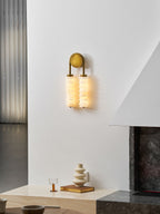 Lit Clérachic sconce in antique brass high on a white wall by a dark stone fireplace, a cream vase on the ledge below.
