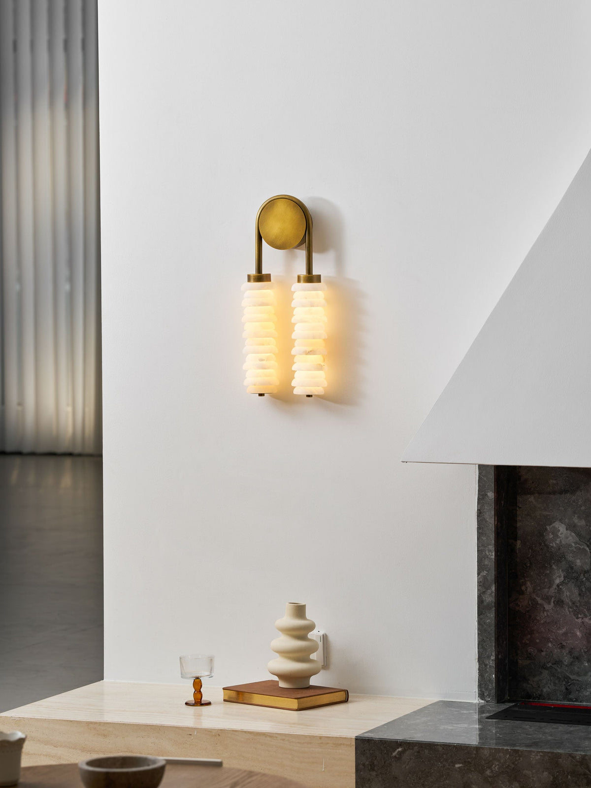 Lit Clérachic sconce in antique brass high on a white wall by a dark stone fireplace, a cream vase on the ledge below.