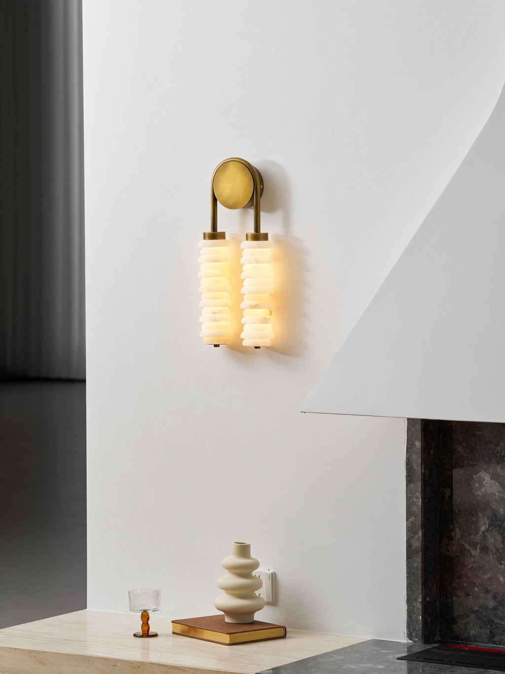 Clérachic sconce in antique brass lit on a white wall, with a grey wall panel on the left and a vase on the hearth below.