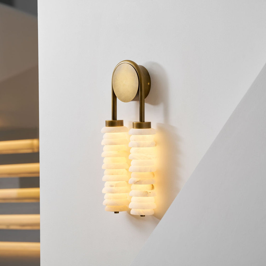 Close-up angle on a lit Clérachic sconce in antique brass, with glowing alabaster discs and a pale round backplate.