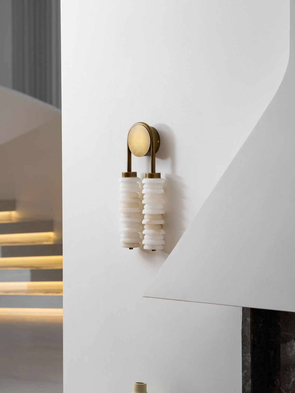 Clérachic sconce in antique brass on a curved white wall, the alabaster columns unlit, with glowing steps in the background.