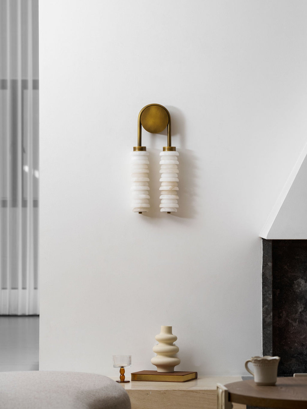 Unlit Clérachic sconce in antique brass on a white wall, above a ledge with a cream vase, a glass and a mug.