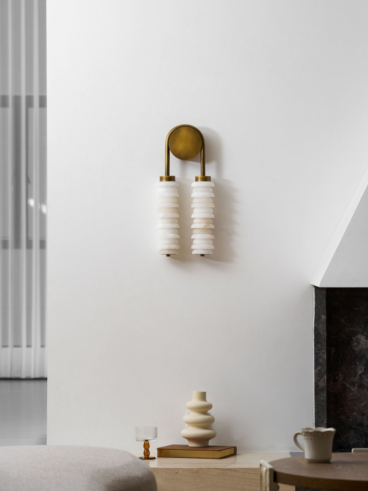 Unlit Clérachic sconce in antique brass on a white wall, above a ledge with a cream vase, a glass and a mug.