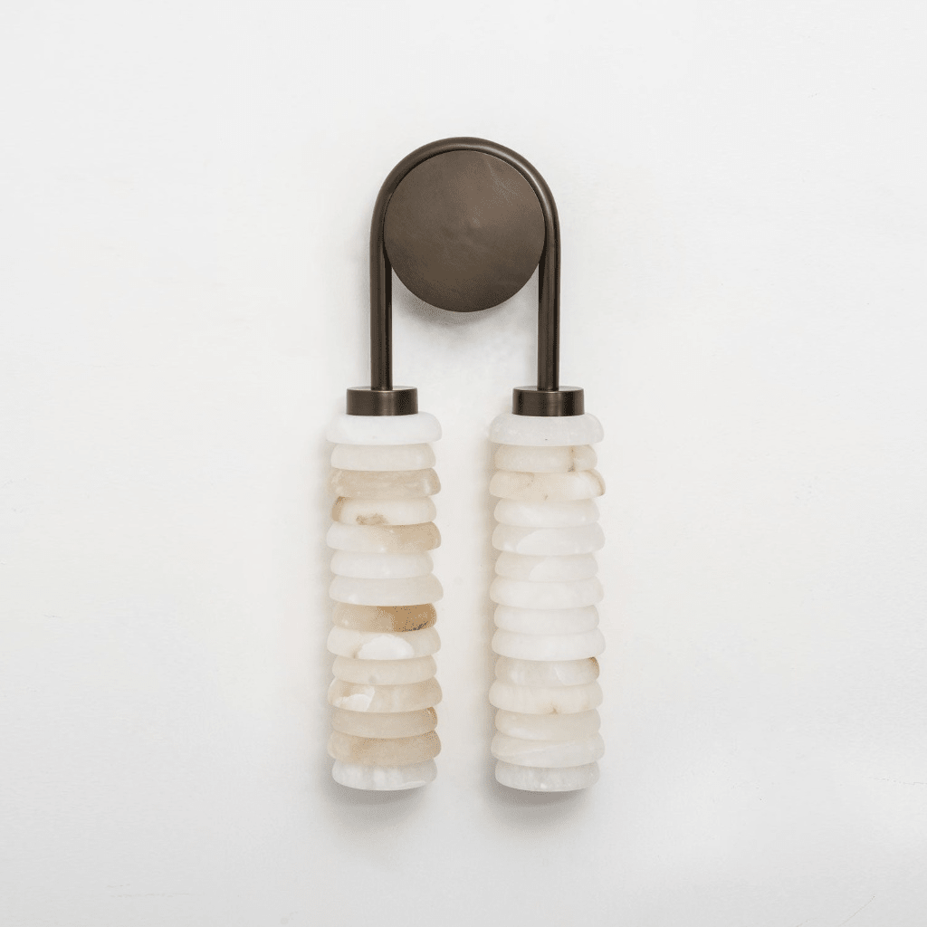 Clérachic sconce in bronze on a white backdrop, with two columns of cream alabaster discs and an arched bronze arm.