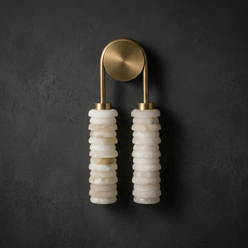 Clérachic sconce in antique brass on a dark charcoal plaster wall, its pale alabaster columns hanging beneath a gold-toned arch.