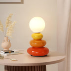 Lit globe of a Clérachic table lamp on a pale pink round table, beside a cream vase of dried pampas grass and a stack of books.