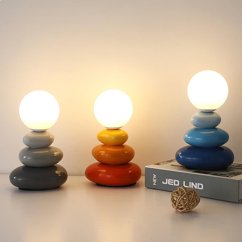 Three Clérachic table lamps side by side, lit, in grey, red-orange and blue, with a hardback book and a woven rattan ball on the shelf.