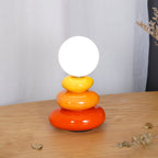 Clérachic stacked-pebble lamp with its globe glowing white, on a wooden table in front of a sheer white curtain, with dried leaves scattered about.