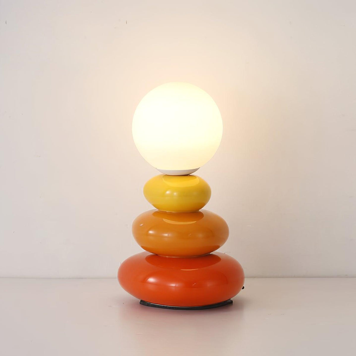 A Clérachic table lamp lit warm on a white surface against a cream wall, its yellow, orange and red ceramic pebbles shining.