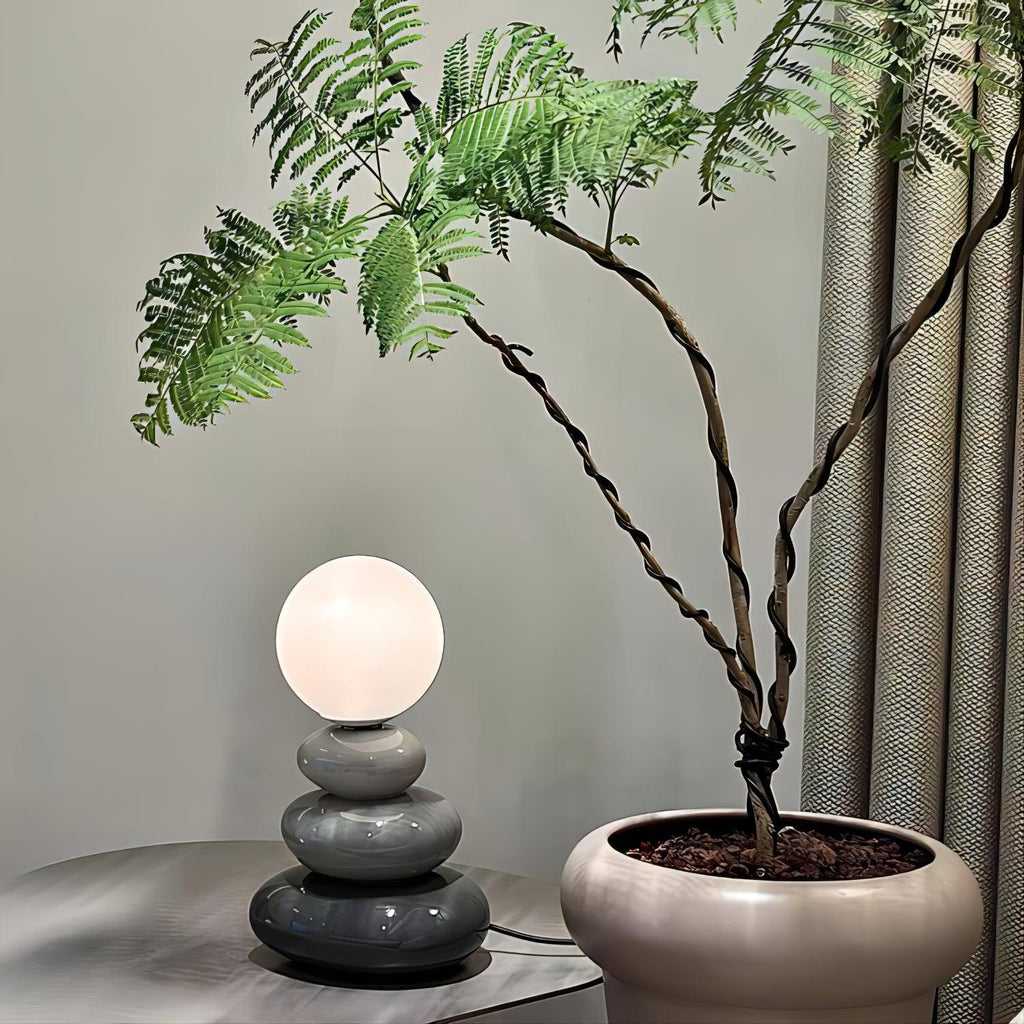 Small grey version of a Clérachic table lamp, lit, on a round metal side table beside a large potted plant with twisted stems.