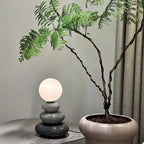 Small grey version of a Clérachic table lamp, lit, on a round metal side table beside a large potted plant with twisted stems.