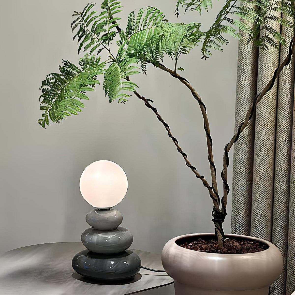 Small grey version of a Clérachic table lamp, lit, on a round metal side table beside a large potted plant with twisted stems.