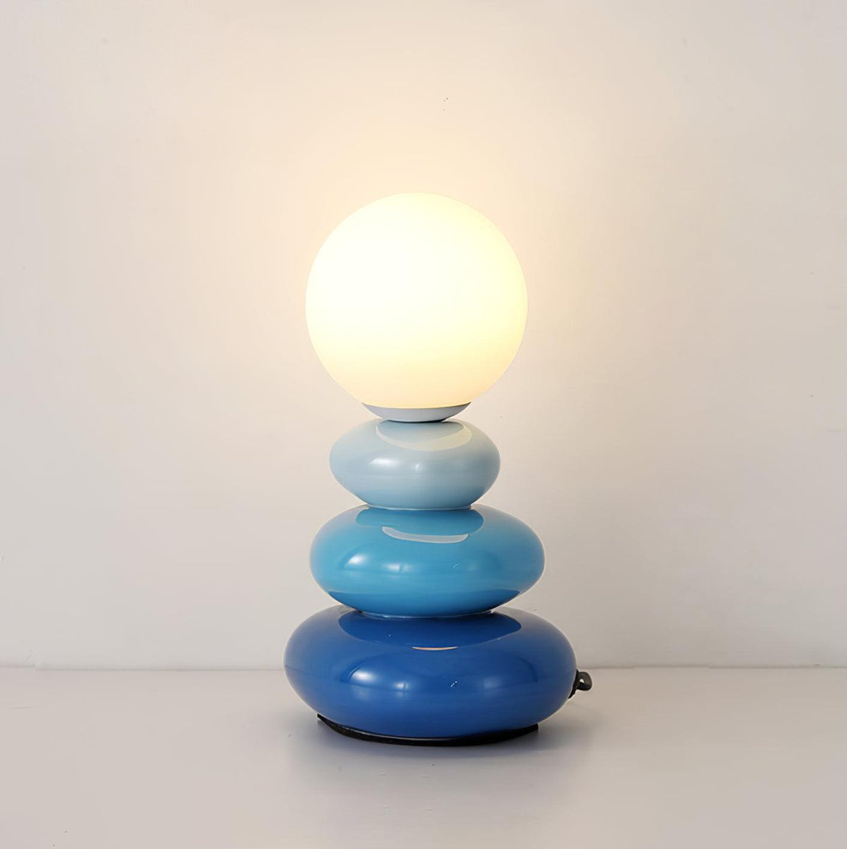 Lit Clérachic table lamp in three shades of blue, from pale to deep, with a frosted globe against a plain white wall.