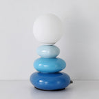 This Clérachic table lamp unlit, a matte white globe over three glossy blue ceramic pebbles on a white tabletop.
