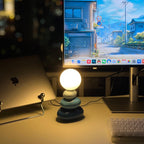 Dark teal-grey version of a Clérachic table lamp, lit warm on a desk beside a laptop, monitor and keyboard.