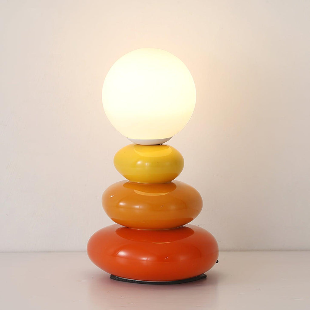 Straight-on view of a Clérachic table lamp, its glowing globe above yellow, orange and red pebbles on a white shelf.