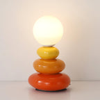 Straight-on view of a Clérachic table lamp, its glowing globe above yellow, orange and red pebbles on a white shelf.