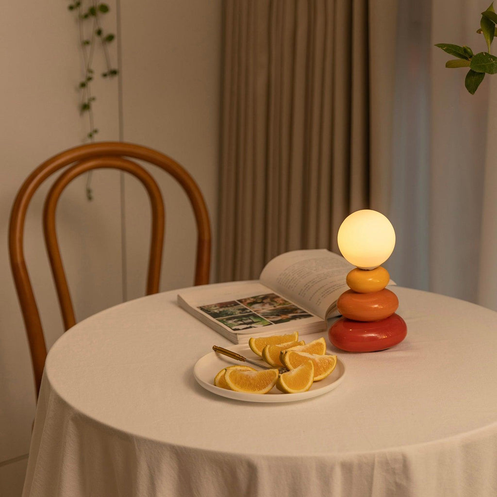 Small Clérachic table lamp lit on a round table with a white cloth, next to an open book and a plate of orange slices, with a bentwood chair behind.