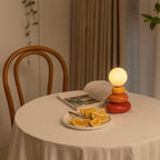 Small Clérachic table lamp lit on a round table with a white cloth, next to an open book and a plate of orange slices, with a bentwood chair behind.