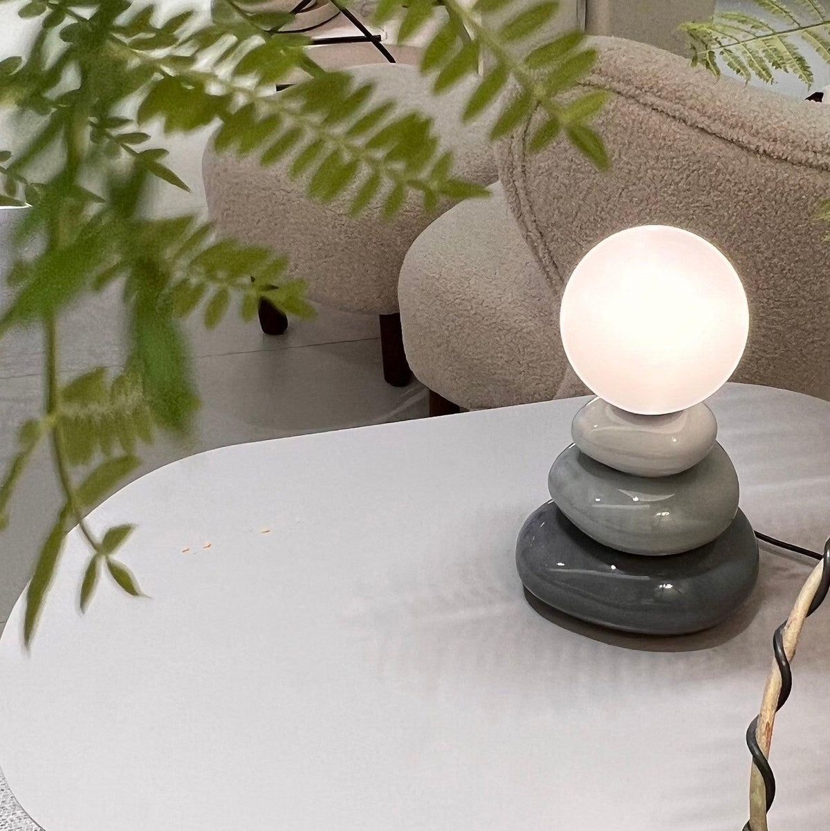 Grey version of a Clérachic table lamp, lit, on a white table with a cream armchair and trailing plant leaves behind it.