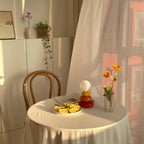 Distant view of a Clérachic table lamp on a small clothed table with orange slices, a vase of poppies and a bentwood chair.