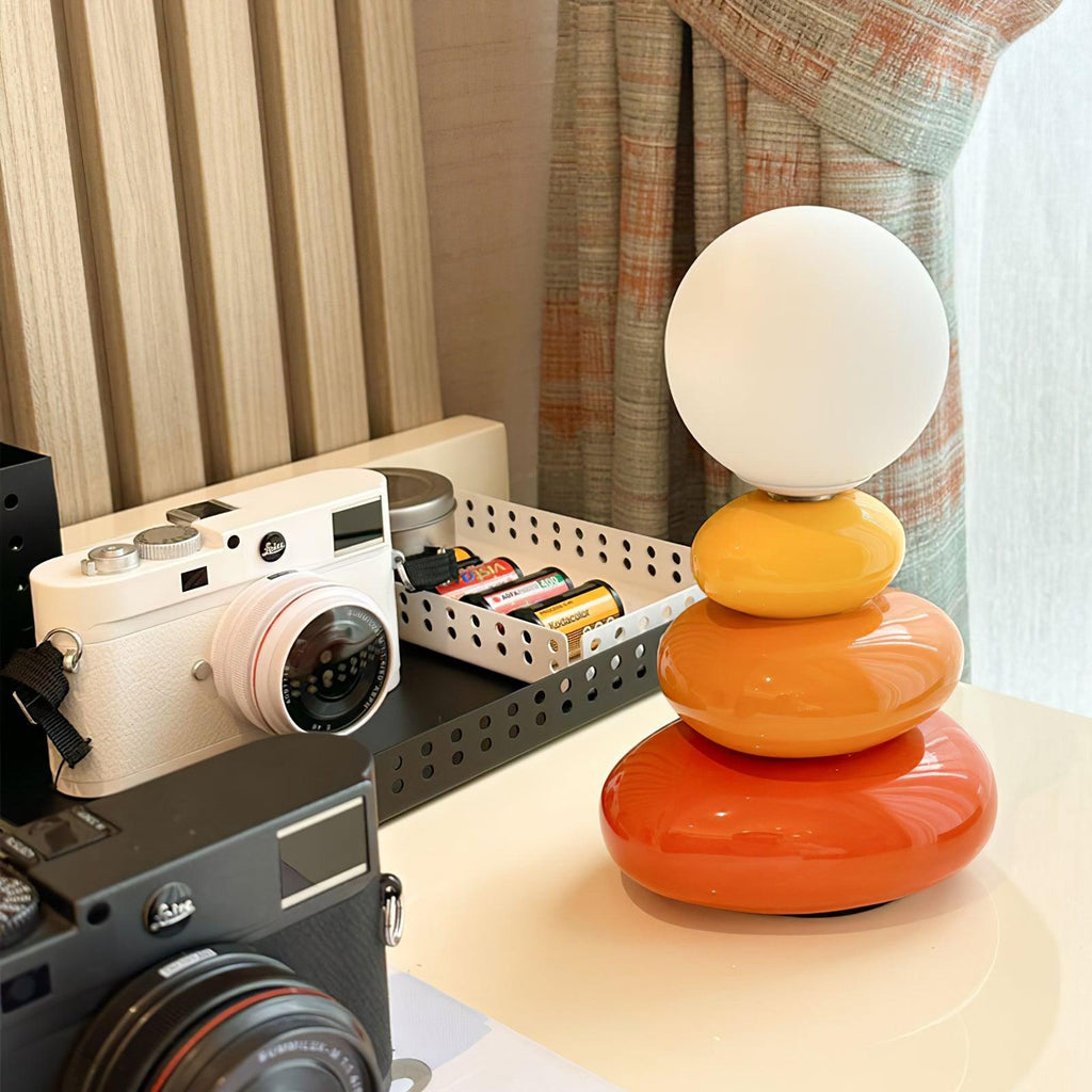 This Clérachic table lamp on a cream desk beside two cameras and a tray of batteries, a striped curtain behind.