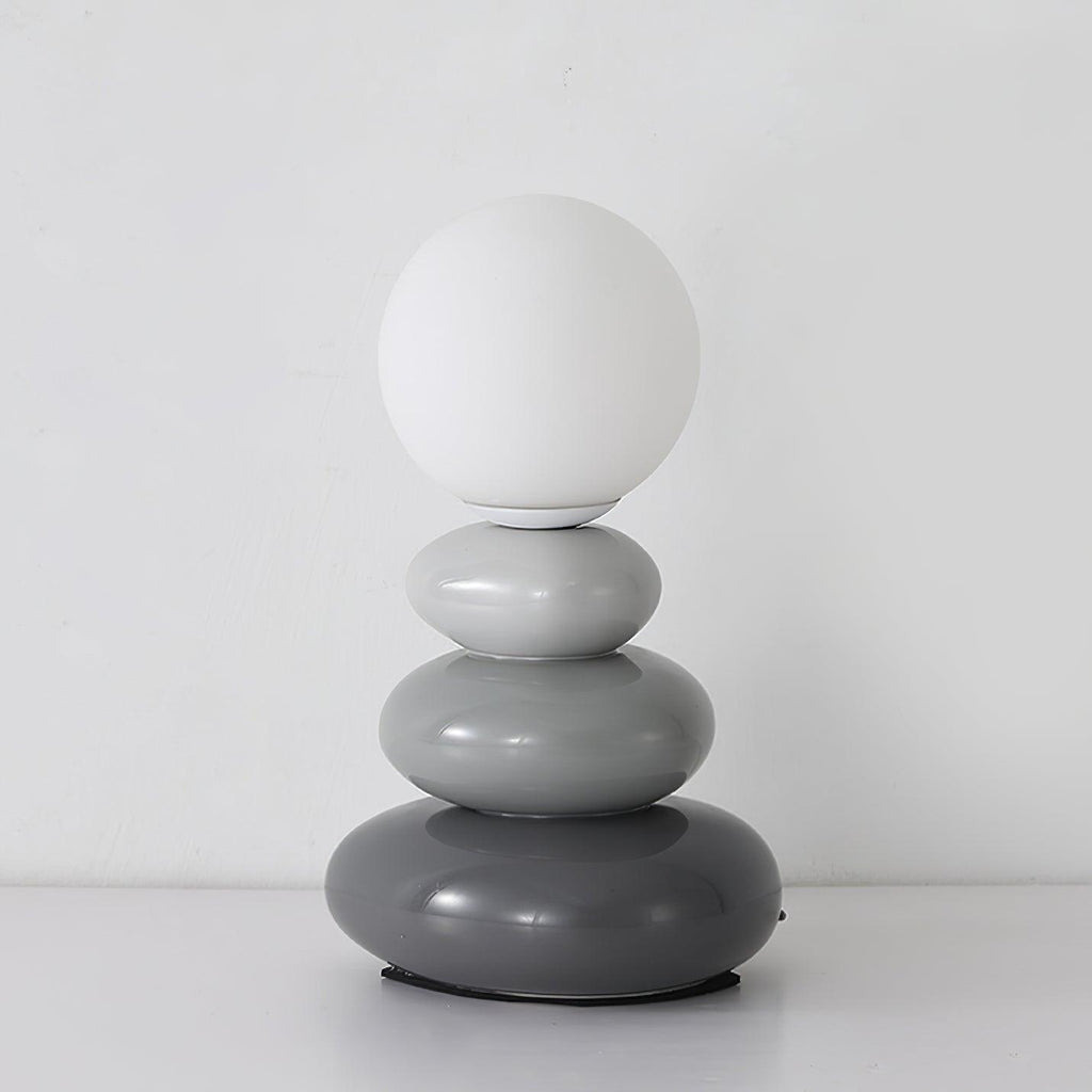 Pale grey to charcoal pebbles under a matte white globe, this Clérachic table lamp unlit on a white surface.