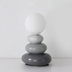 Pale grey to charcoal pebbles under a matte white globe, this Clérachic table lamp unlit on a white surface.