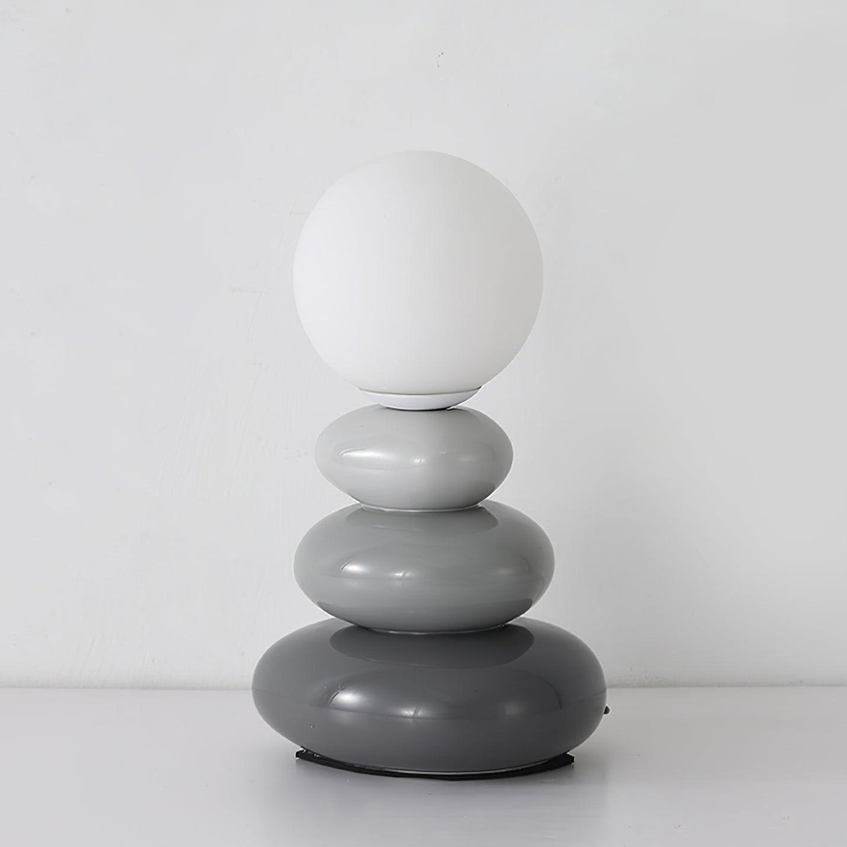 Pale grey to charcoal pebbles under a matte white globe, this Clérachic table lamp unlit on a white surface.