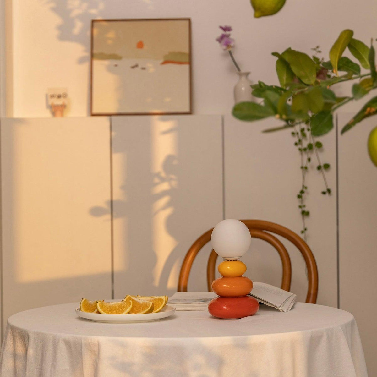 A Clérachic table lamp on a clothed round table by a bentwood chair, with orange slices and a framed print on the wall behind.