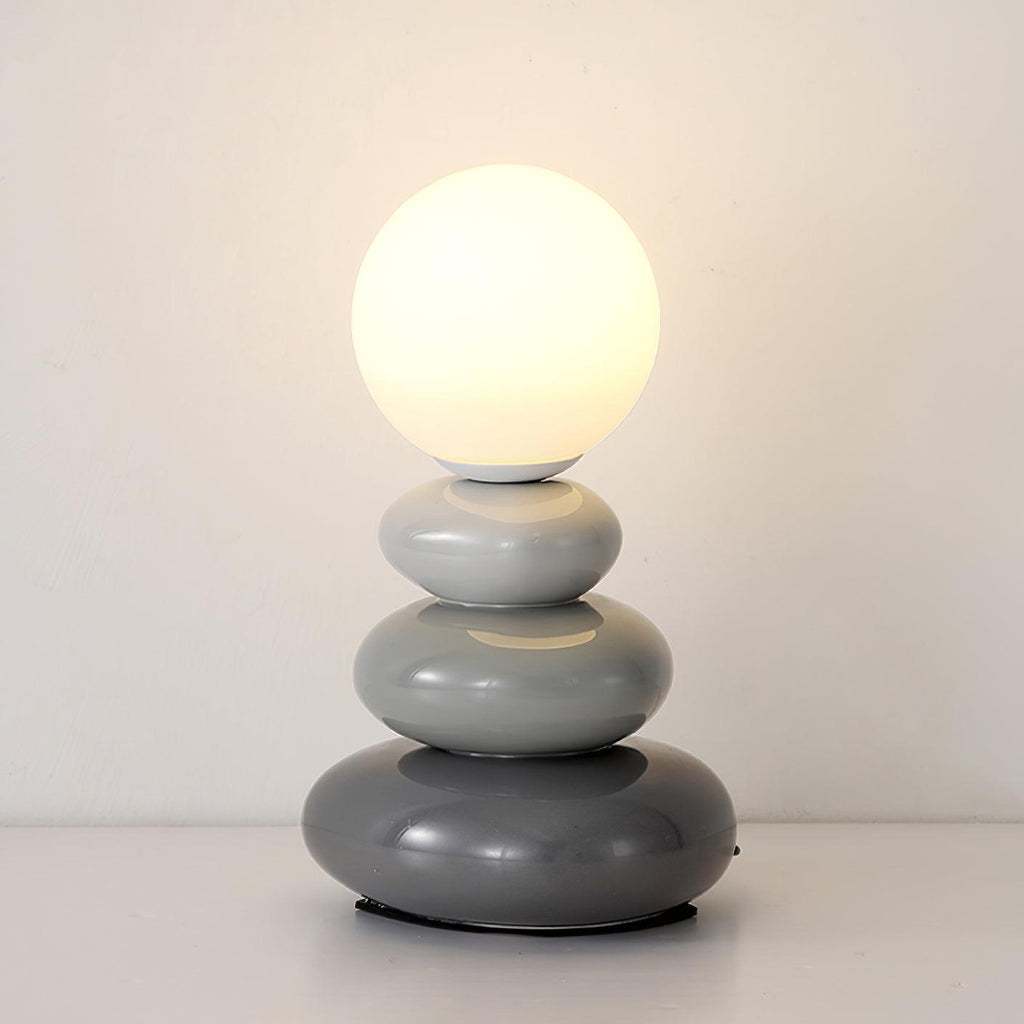 Lit Clérachic table lamp with three grey pebbles, light to dark, under a warm glowing globe against a white wall.