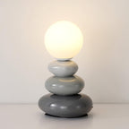 Lit Clérachic table lamp with three grey pebbles, light to dark, under a warm glowing globe against a white wall.
