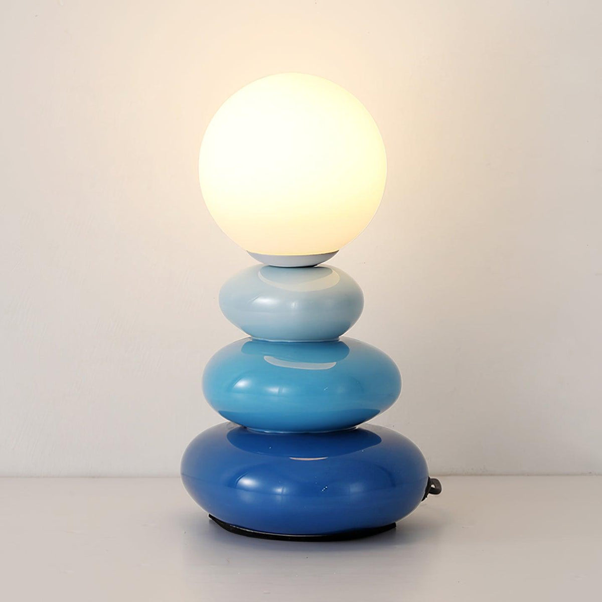 Blue Clérachic table lamp, lit, its pebbles shading from pale sky blue to deep cobalt beneath a warm white globe.