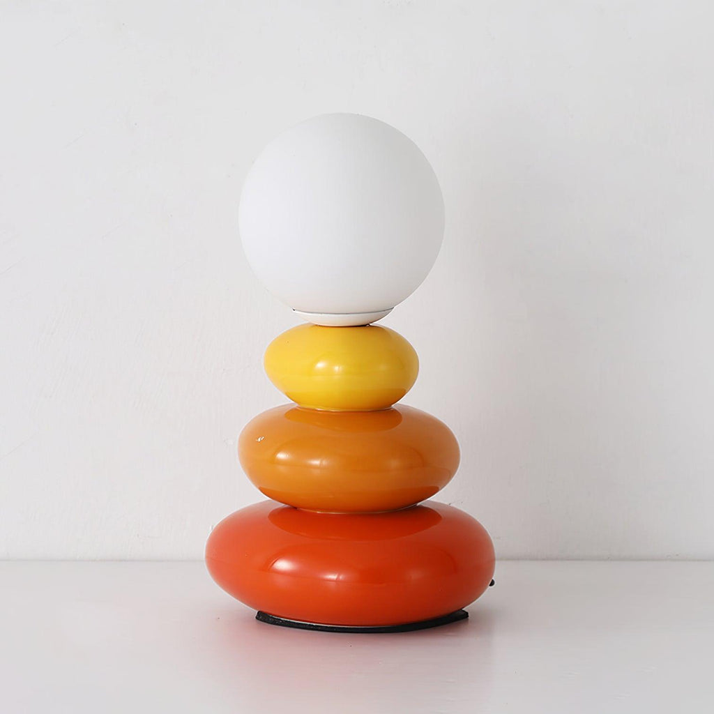 This Clérachic table lamp unlit on a white shelf, a matte globe over yellow, orange and red glossy pebbles.