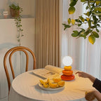 Clérachic table lamp lit on a small round table with an open book and a plate of orange slices, a hand reaching in at the right.