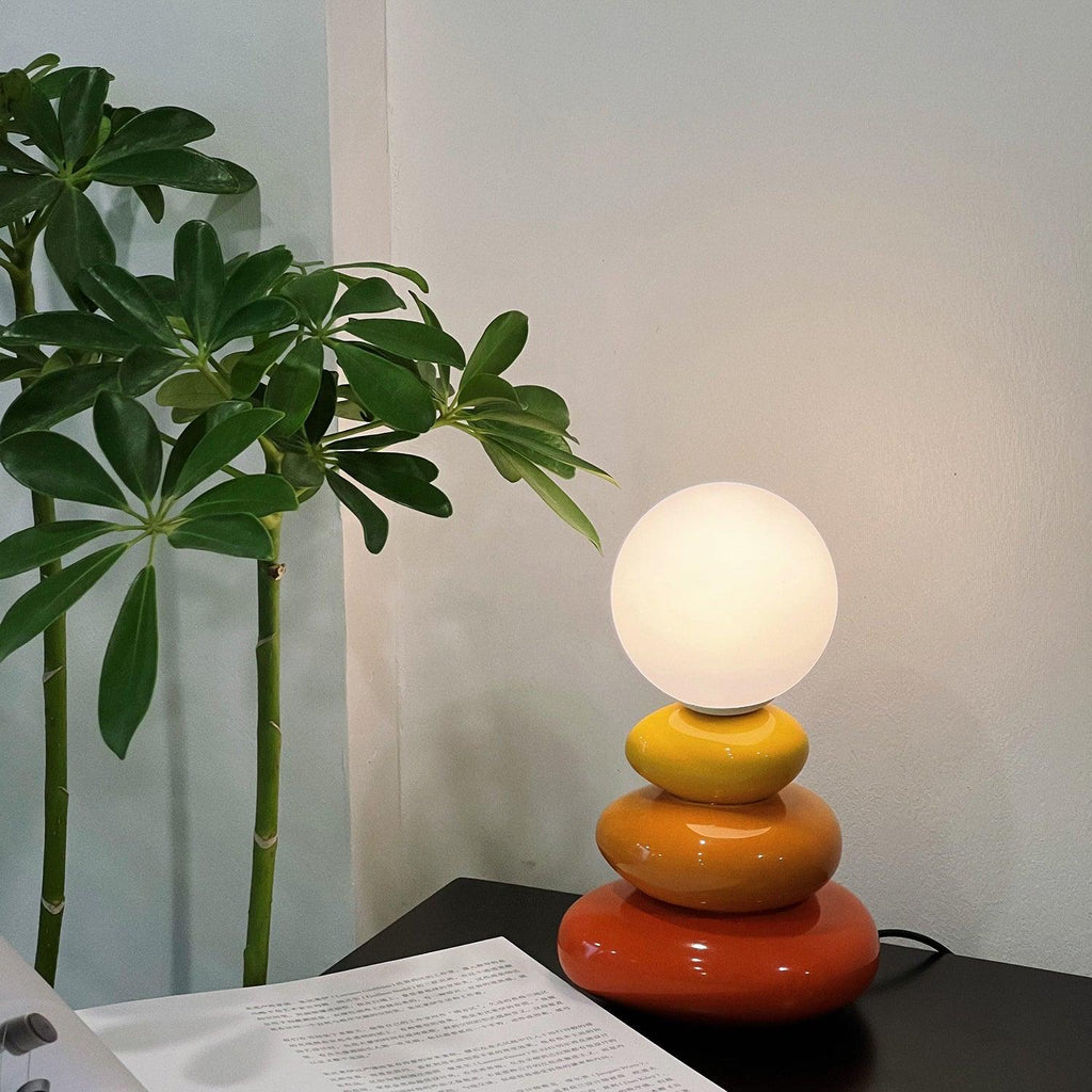 A Clérachic table lamp glowing on a dark desk beside a tall green plant, with printed pages in the foreground.