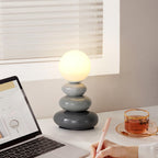 Grey Clérachic table lamp lit beside a window blind, on a white desk with an open laptop, a cup of tea and a hand holding a pen.