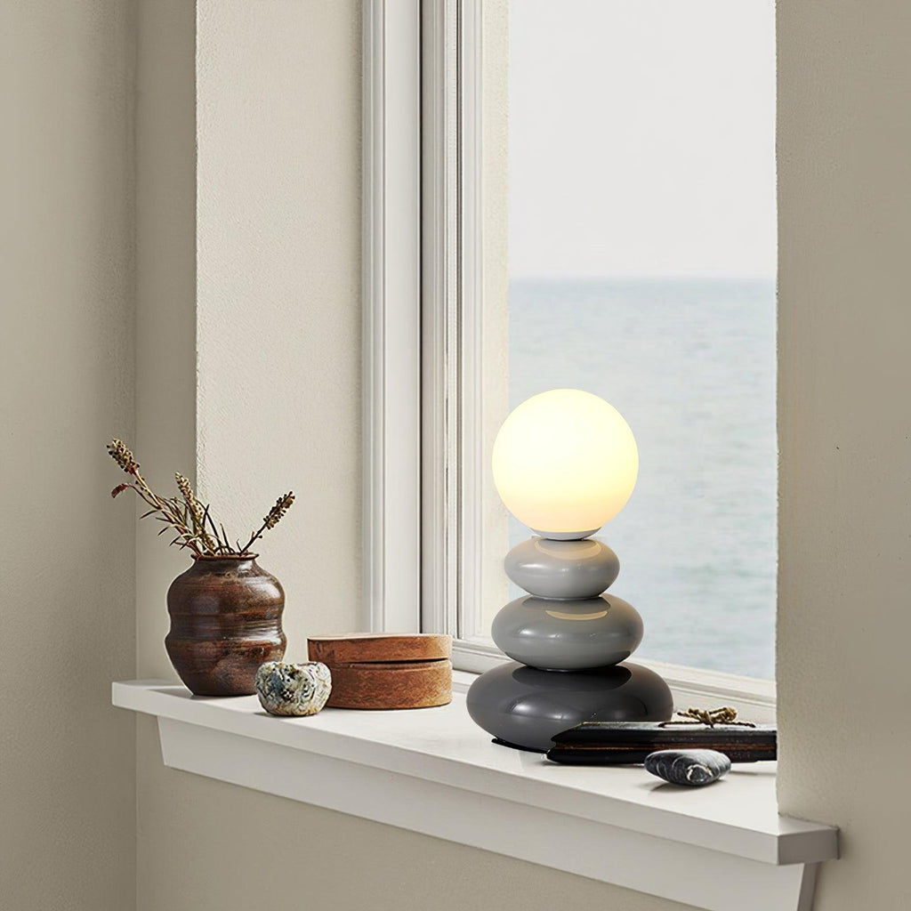 A grey Clérachic table lamp lit on a white window sill, beside a brown ceramic vase with twigs and a view of the sea.