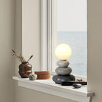 A grey Clérachic table lamp lit on a white window sill, beside a brown ceramic vase with twigs and a view of the sea.