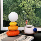 Clérachic table lamp on a magazine in front of a green leafy print, with a white cup of coffee and a blue ball handle beside it.