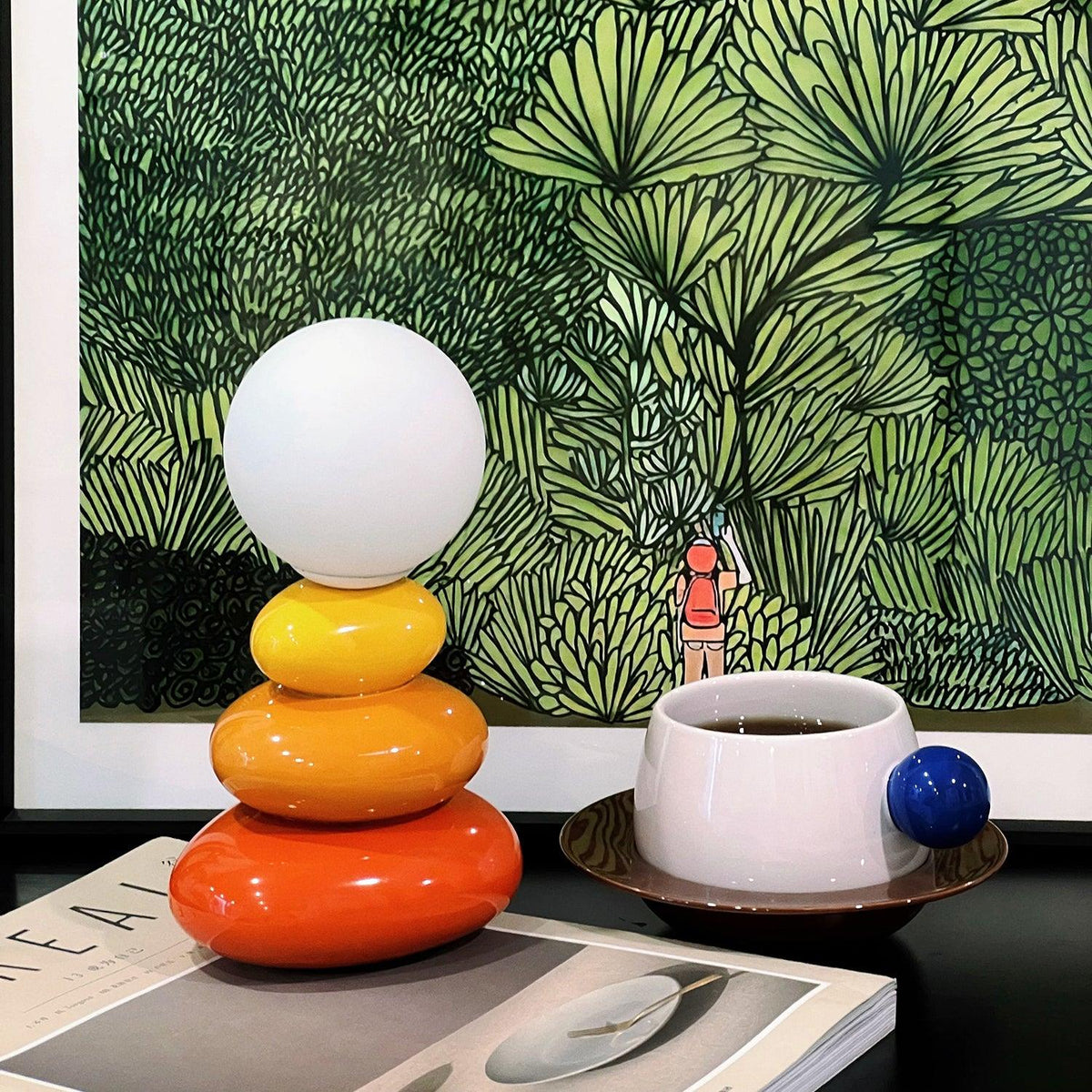 Clérachic table lamp on a magazine in front of a green leafy print, with a white cup of coffee and a blue ball handle beside it.