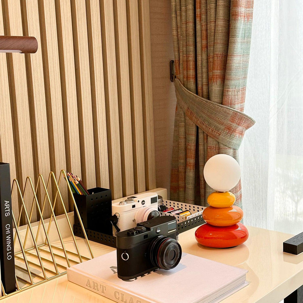 Clérachic table lamp on a cream desk with a camera, a gold file holder and a book, patterned curtain at the window.