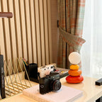 Clérachic table lamp on a cream desk with a camera, a gold file holder and a book, patterned curtain at the window.