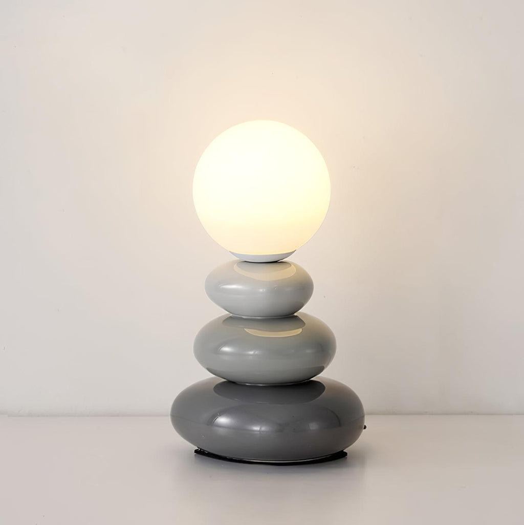 Grey Clérachic table lamp lit warm against a plain wall, three smooth ceramic pebbles in graduated grey under a glass globe.