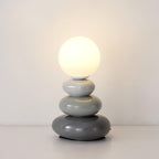 Grey Clérachic table lamp lit warm against a plain wall, three smooth ceramic pebbles in graduated grey under a glass globe.