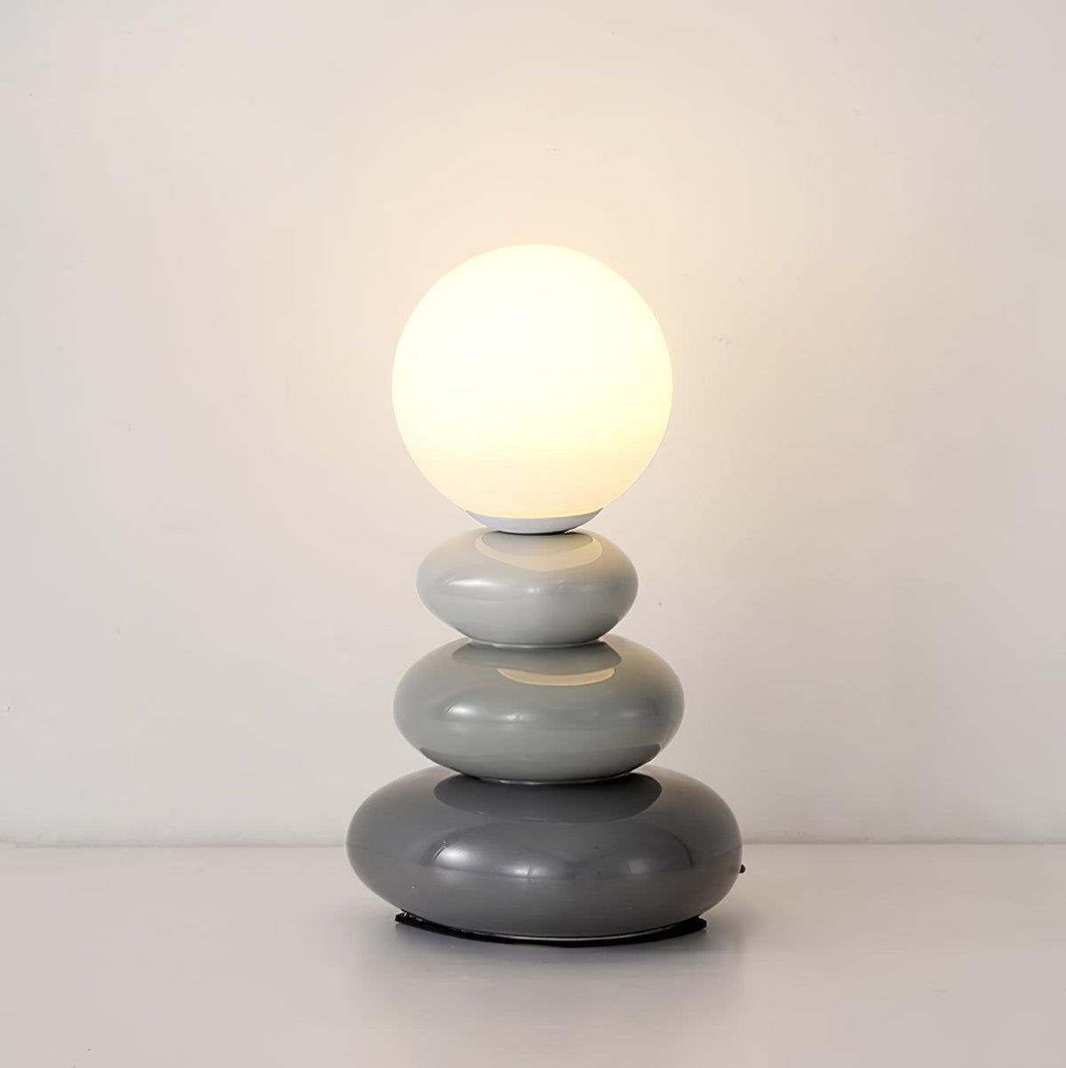 Grey Clérachic table lamp lit warm against a plain wall, three smooth ceramic pebbles in graduated grey under a glass globe.