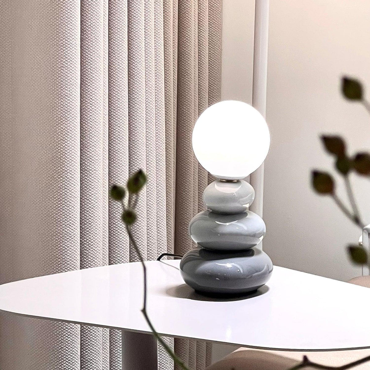Lit grey Clérachic table lamp on a white table with a pale curtain behind and a sprig of dark leaves in the foreground.
