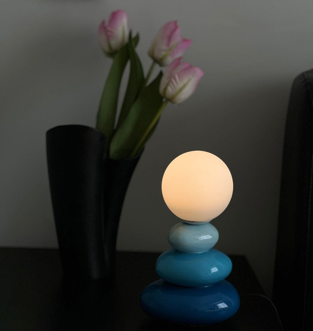 Blue Clérachic table lamp lit in a dim room, with a black vase of pink tulips beside it.